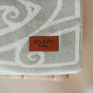 fern coloured patterned blanket with a brown leather label reading 'BO&KO baby' on a blurred background.
