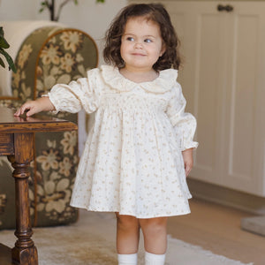 Young girl in a white dress standing in a room with a wooden table and floral-patterned chair.