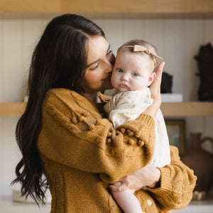 Woman in a mustard yellow sweater holding a baby in a white outfit with a bow.