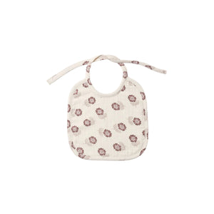 A cream-colored muslin baby bib with a small brown and pink floral pattern, featuring tie straps at the top, shown against a plain white background.